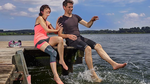 Couple sitting on a landing stage dangling joyfully their feet in a lake while showcasing a medi knee brace